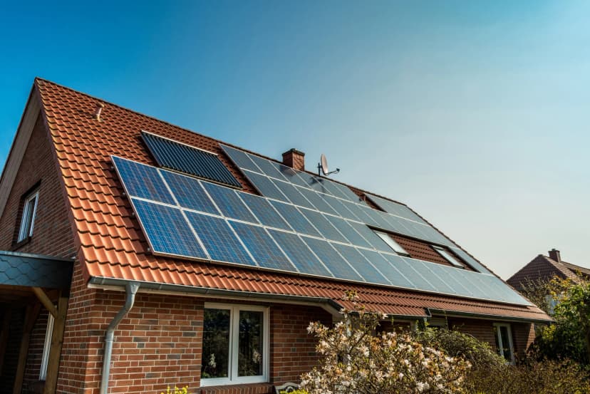 The sun is shining on four rows of solar panels on a red-roofed house.