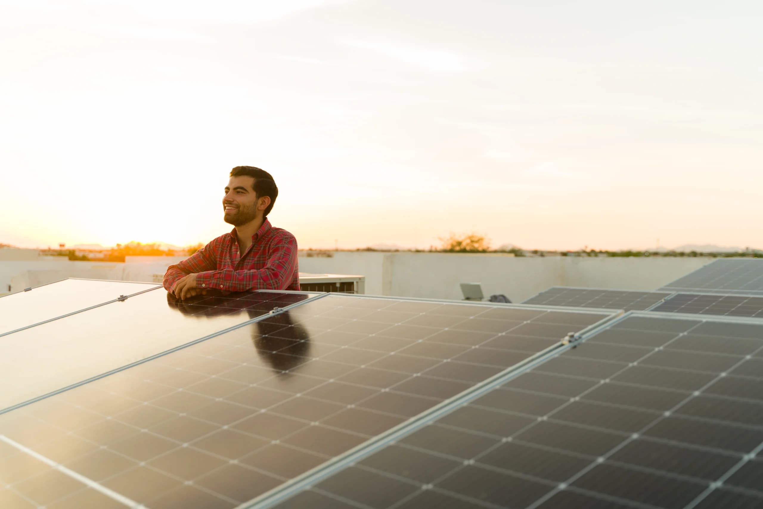 A man is standing on his flat roof, resting his arms on his newly installed solar panels.